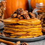 stack of pumpkin pancakes with candied pecans on top and syrup being drizzled onto pancakes with pumpkins in the background