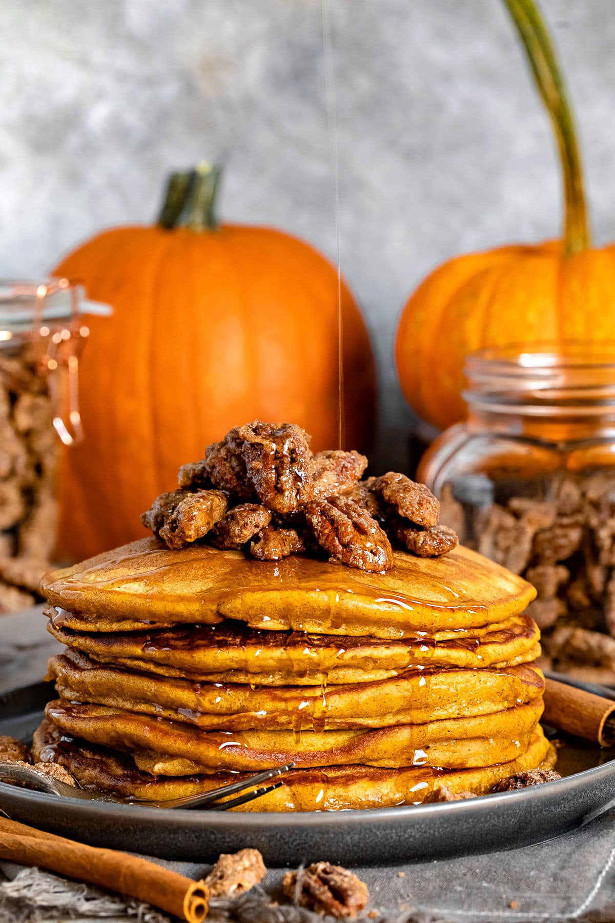 stack of pumpkin pancakes with candied pecans on top and syrup being drizzled onto pancakes with pumpkins in the background