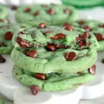 Two mint chocolate chip cookies with one half cookie on top with melty chocolate. Cookies are green with chocolate chips. Green cake stand can be seen in the background.