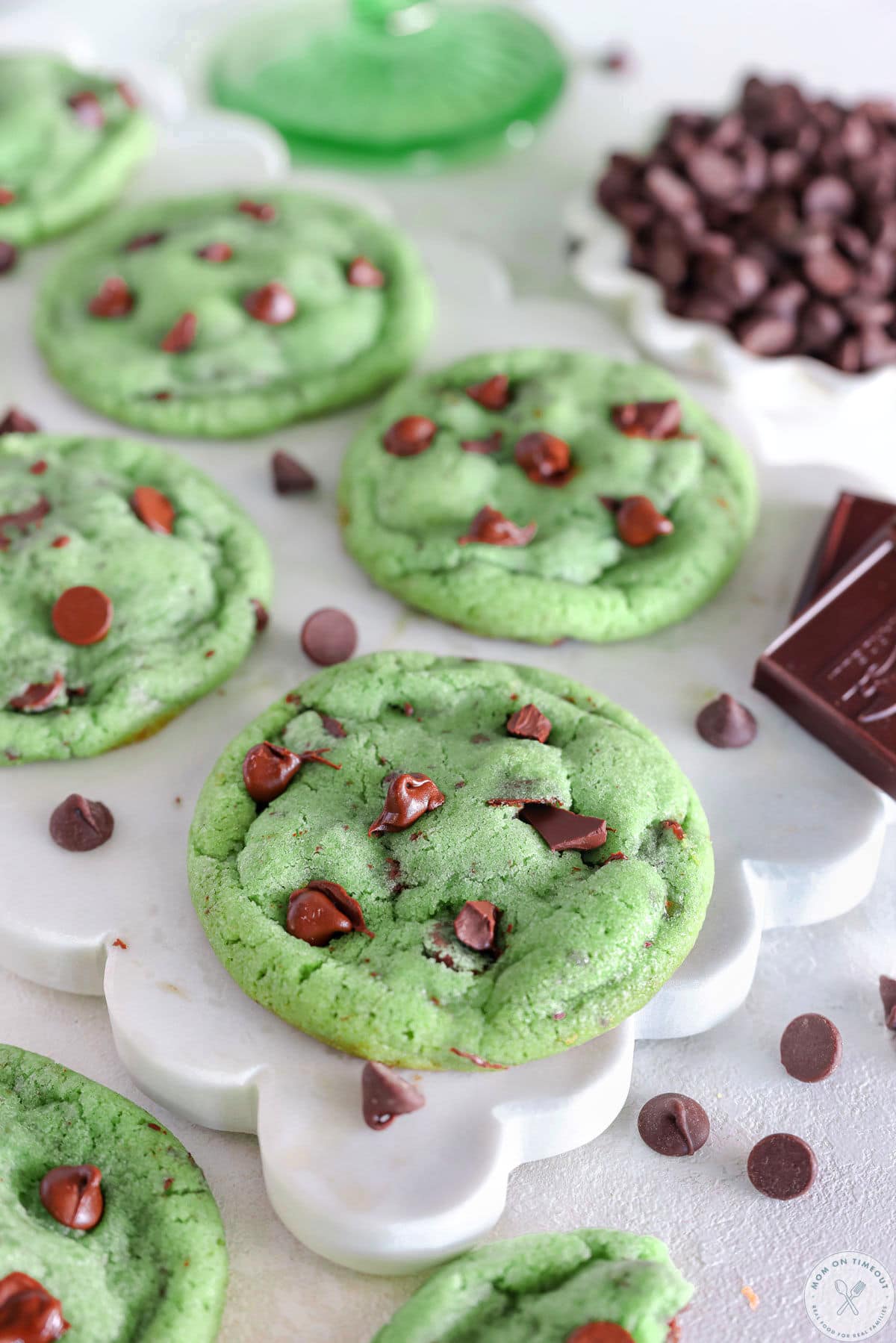 Four mint chocolate chip cookies baked and resting on white marble board with bowl of chocolate chips next to it. 