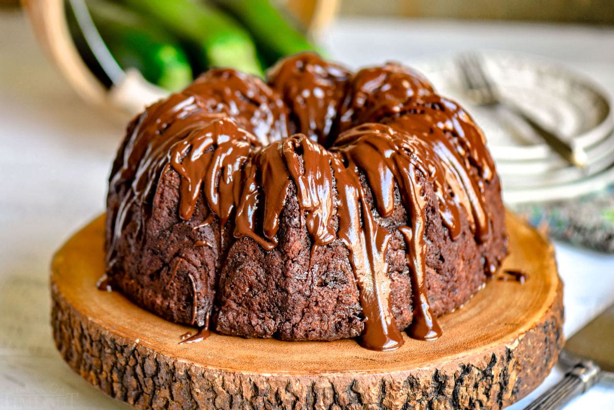 chocolate zucchini cake made in a bundt pan on wood cake stand with zucchinis in the background.