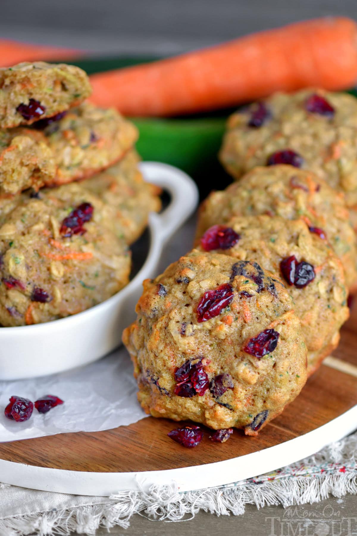 Zucchini oatmeal cookies in a row on a wood board. Craisins can be seen on top of the cookies along with a carrot and zucchini in the background.