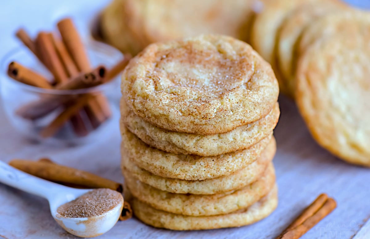 snickerdoodle cookies stacked on cutting board with cookies in background and cinnamon stick scattered around