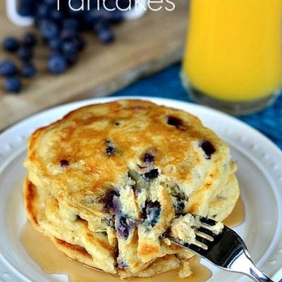 a stack of Blueberry Buttermilk Pancakes on white plate with syrup and a cup of orange juice in the back