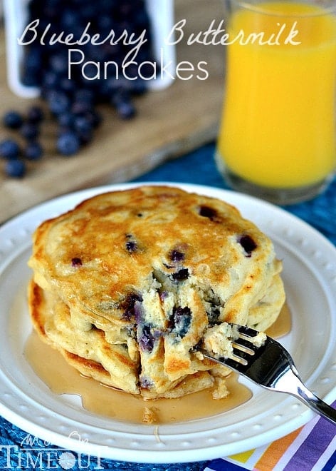 a stack of Blueberry Buttermilk Pancakes on white plate with syrup and a cup of orange juice in the back