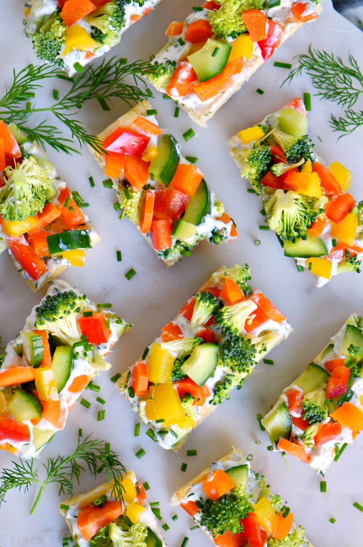 top down view of veggie pizza recipe assemble and cut into small pieces for serving. dill sprigs are scattered about on the marble cutting board.