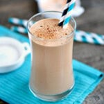 Coffee protein shake in a tall glass with two paper straws. Glass is sitting on teal napkin on a wood surface.