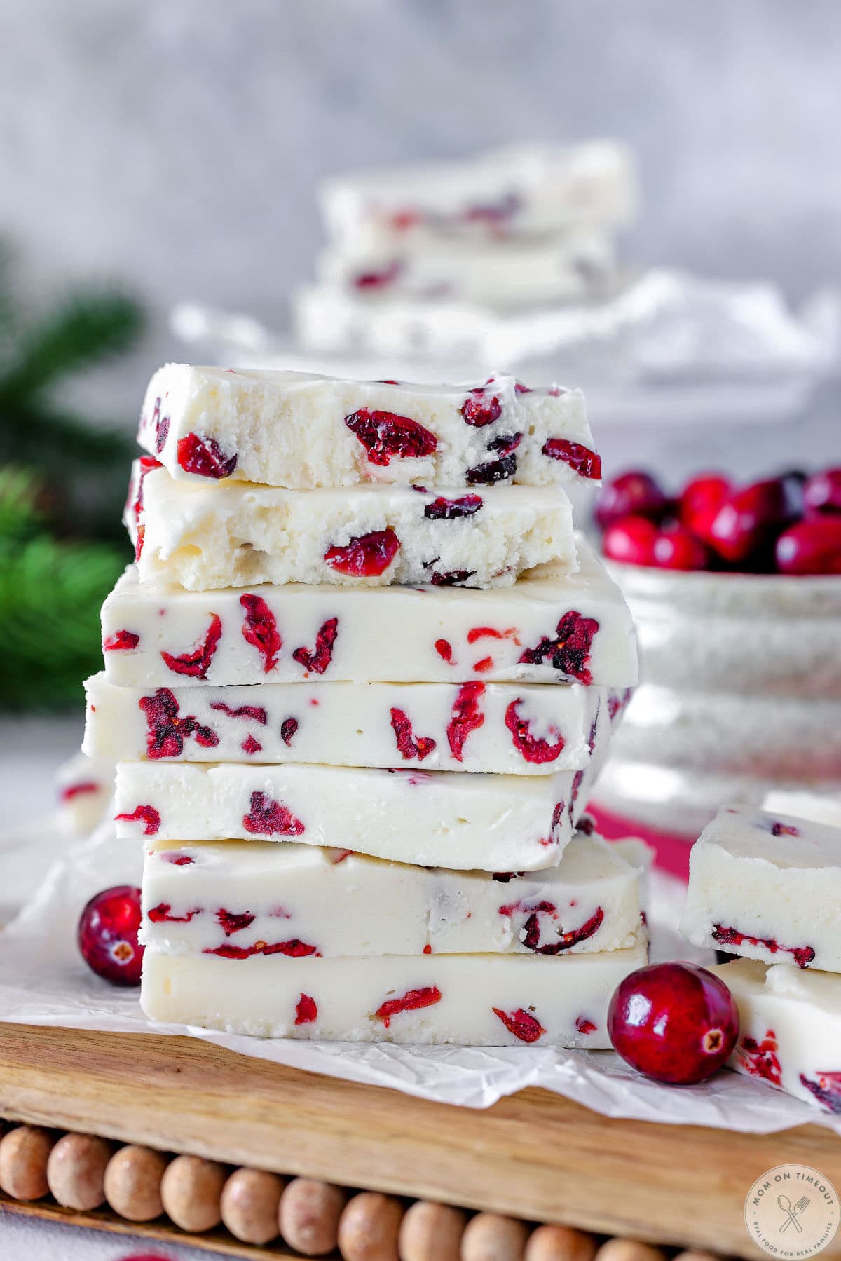 Tall stack of cranberry fudge on wood and marble cutting board. Fresh cranberries and more fudge can be seen in the background.