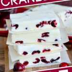 white chocolate cranberry fudge cut into squares and stacked four high sitting on a wood board on a square of parchment paper. Text on diagonal color blocks at the top and bottom of the image.