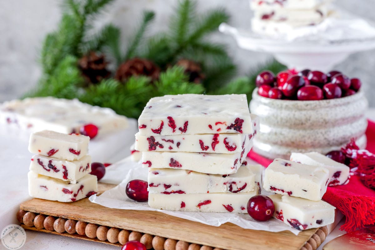 Wide shot of white chocolate cranberry fudge stacked on wood and marble cutting board with fresh cranberries off to the side.