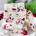 White chocolate cranberry fudge stacked high on vintage white pedestal. Greenery and fresh cranberries can be seen in background.