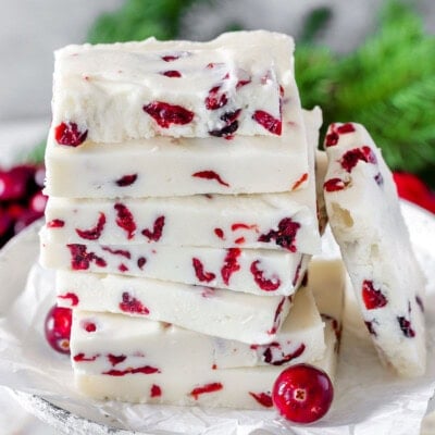 White chocolate cranberry fudge stacked high on vintage white pedestal. Greenery and fresh cranberries can be seen in background.