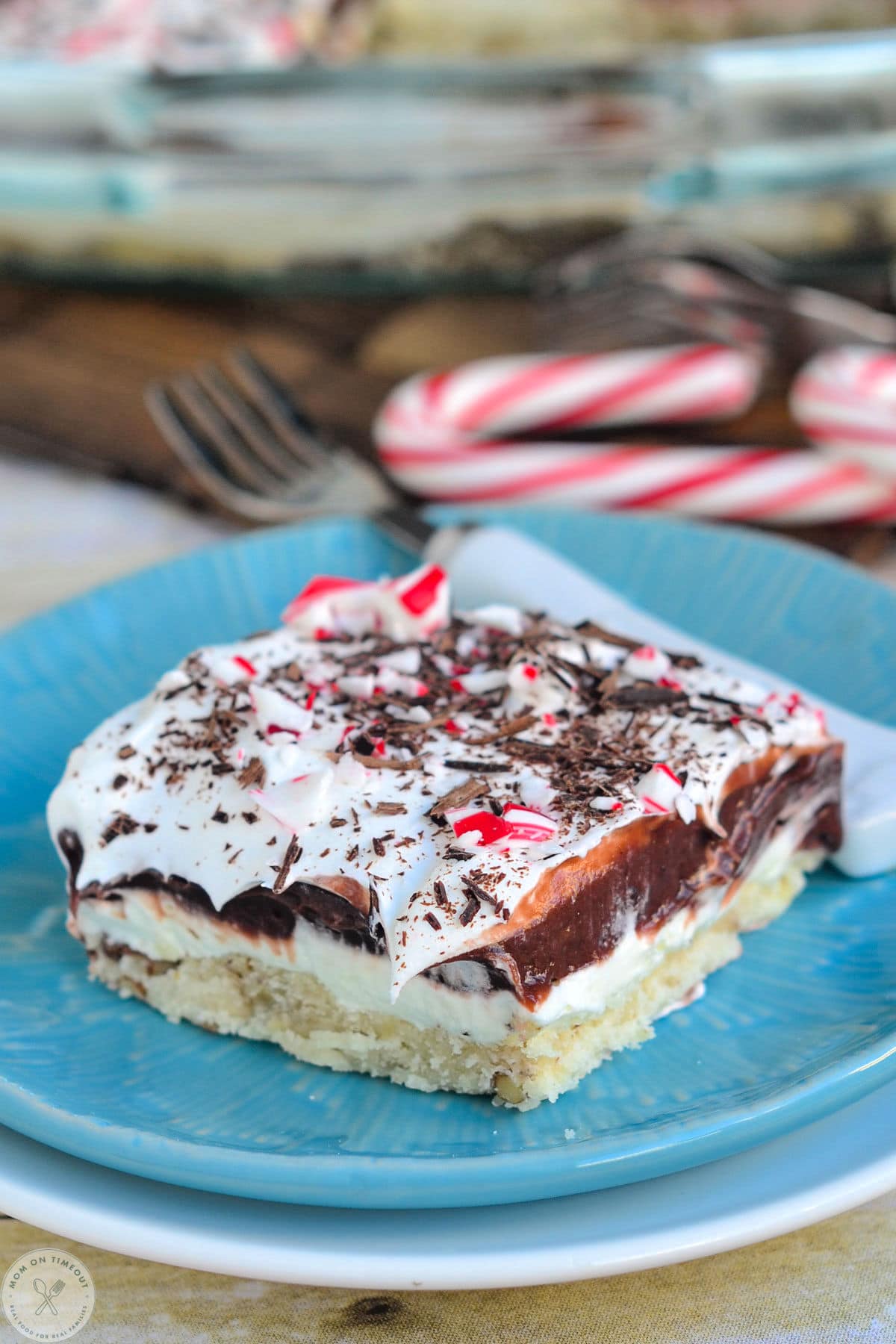 Four layered chocolate peppermint dessert on blue plate topped with candy canes. Chocolate Peppermint Lush is topped with crushed candy canes.