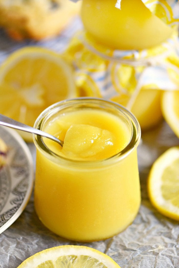 lemon curd in a glass jar with a spoon with sliced lemons around it