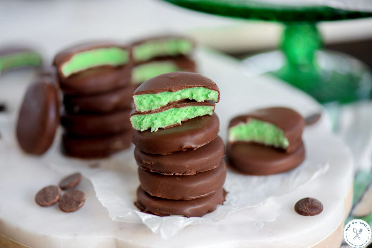 Wide shot of chocolate mint patties on a marble board with three of the patties split in half showing the creamy, green mint interior.