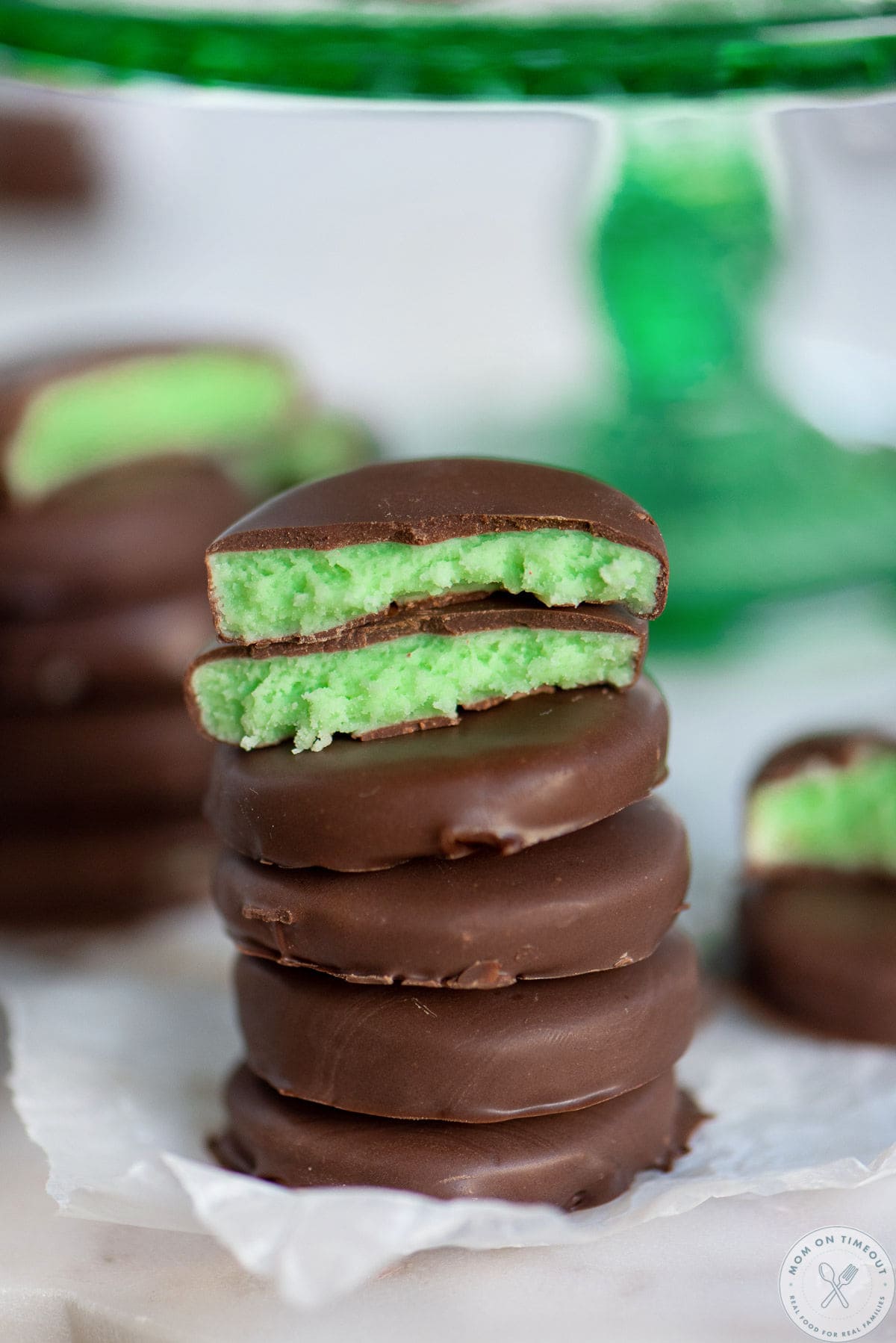 Homemade mint patties stacked five high with the top one split in half. A green cake stand in the background has more patties on it.