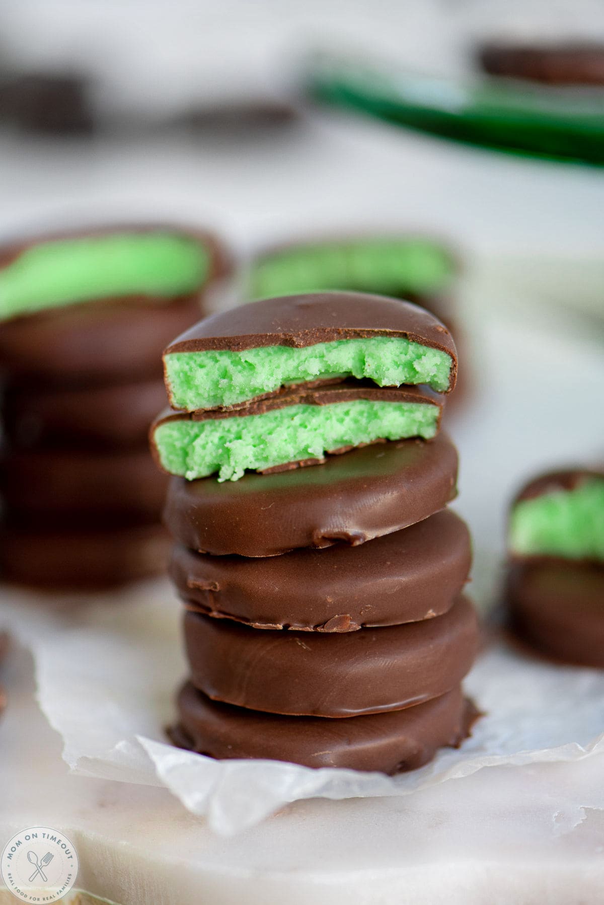 Homemade mint patties stacked five high with the top one split in half. A green cake stand in the background has more patties on it.