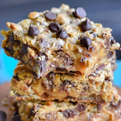Three cookie bars stacked on a wood board. Bars are made with peanut butter, caramel, toffee, chocolate chips and oats.