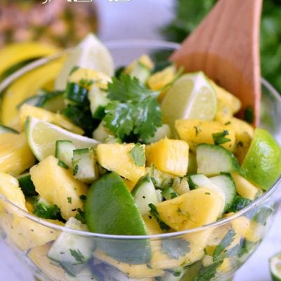 refreshing pineapple cucumber salad in clear plastic bowl with large wood serving spoon. title overlay at top of image.