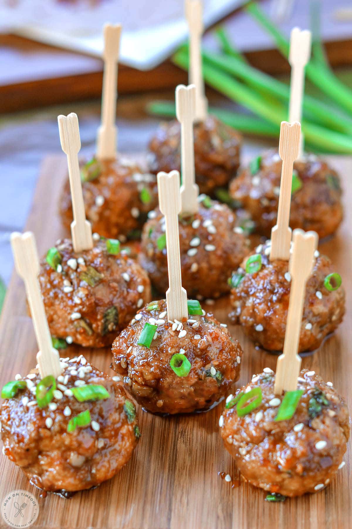 Nine teriyaki meatballs on wood cutting board topped with green onions and sesame seeds. Each meatball has a toothpick in it.