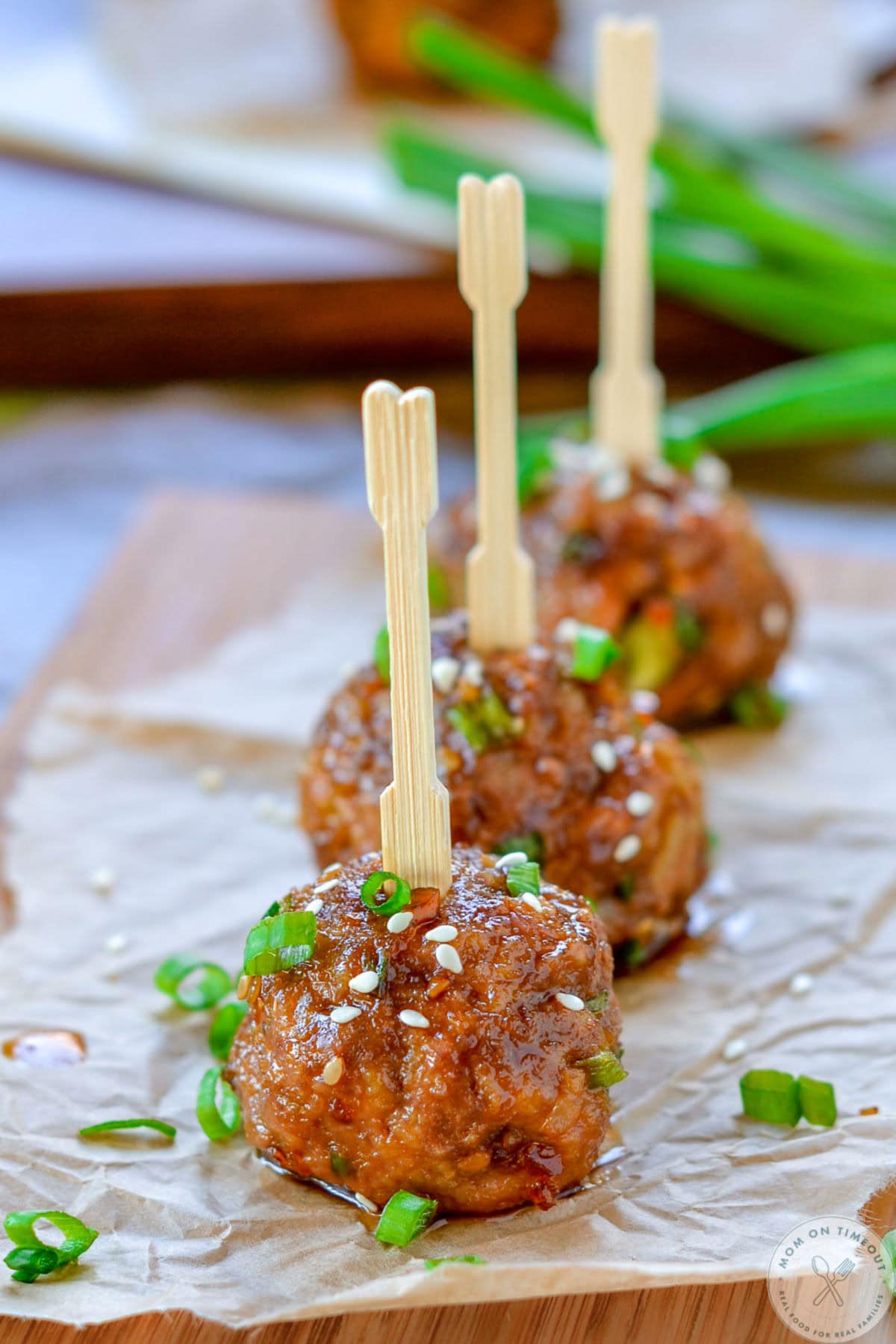 Three teriyaki meatballs lined up on parchment paper, topped with green onions and sesame seeds. Toothpicks have been inserted into the top of each meatball.