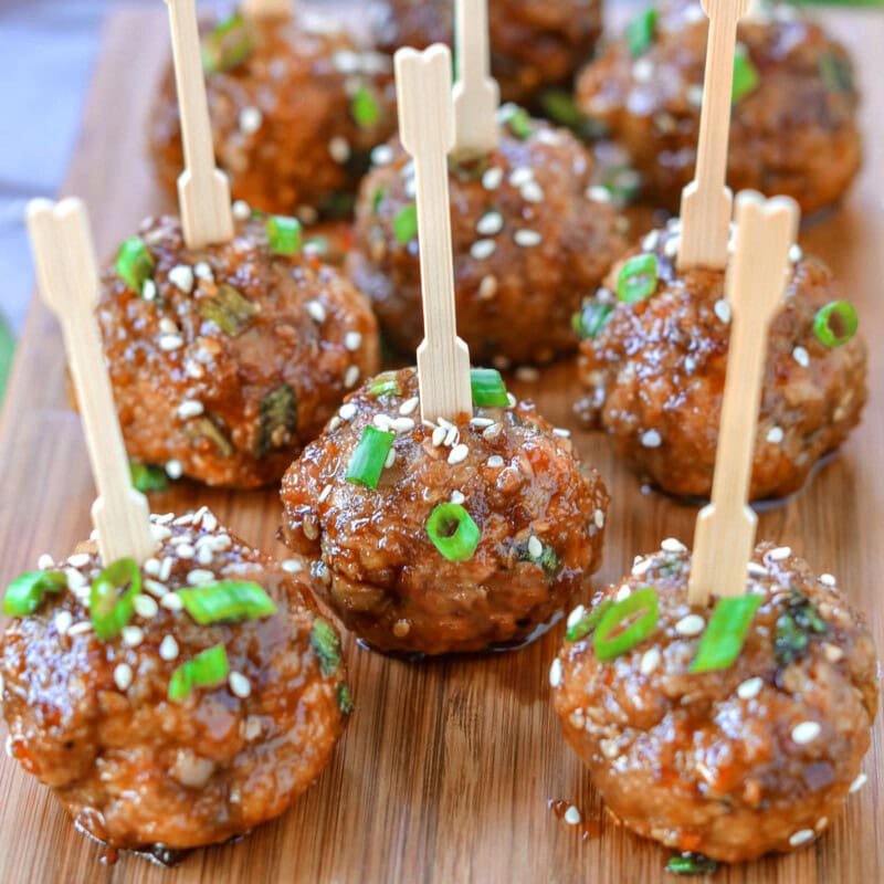 Nine teriyaki meatballs on wood cutting board topped with green onions and sesame seeds. Each meatball has a toothpick in it.