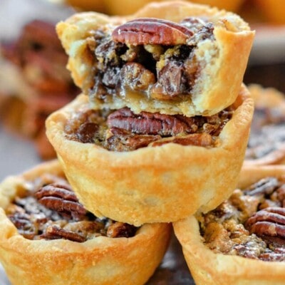 five mini pecan pies stacked in a pyramid with more pies in the background. the top pie has is cut in half. each mini pie is stopped with a pecan half.