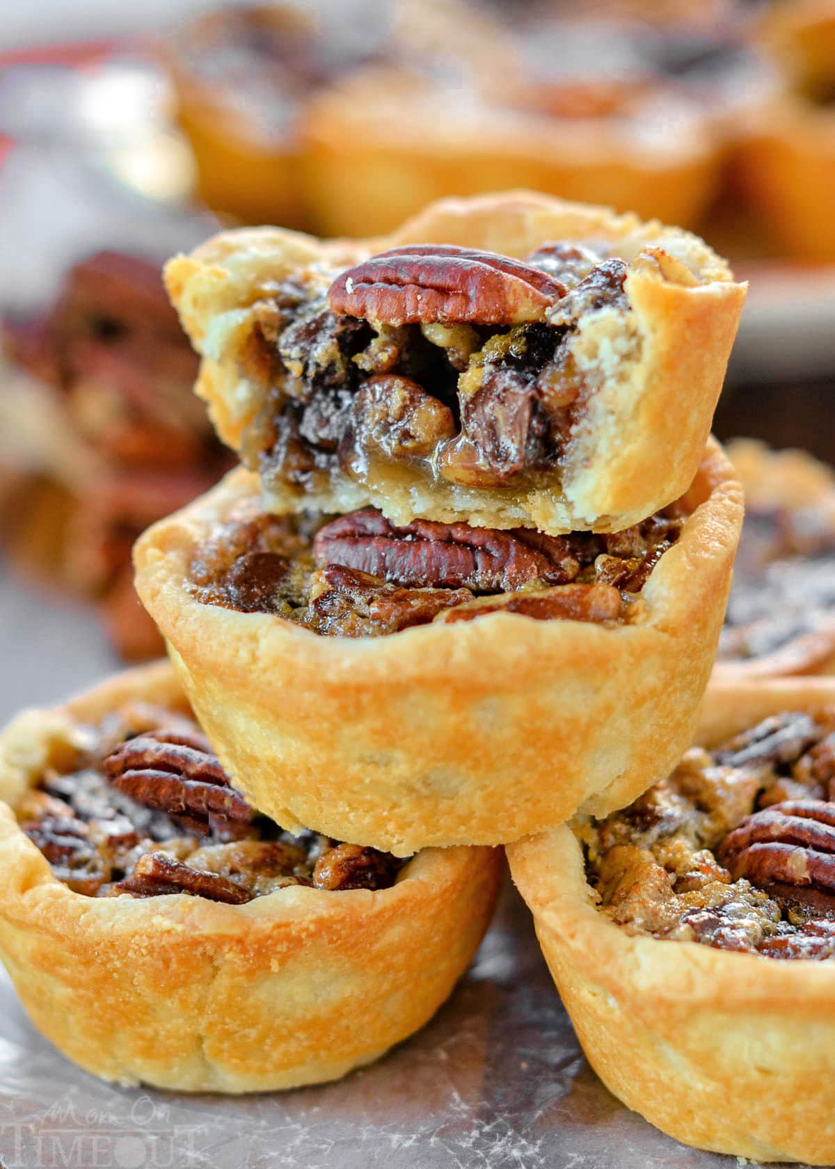 five mini pecan pies stacked in a pyramid with more pies in the background. the top pie has is cut in half. each mini pie is stopped with a pecan half.