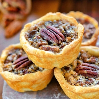 four mini pecan pies stacked in a pyramid with more pies in the background. each mini pie is stopped with a pecan half.
