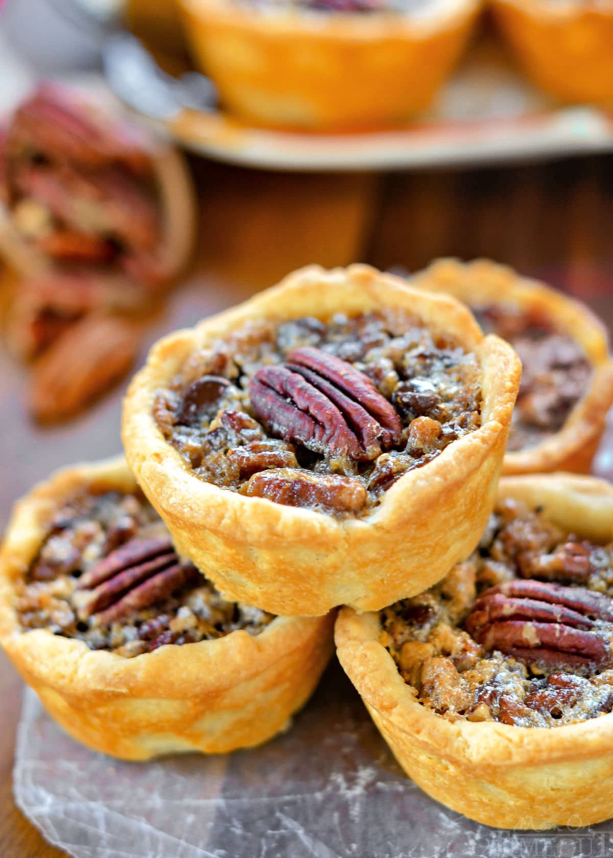 four mini pecan pies stacked in a pyramid with more pies in the background. each mini pie is stopped with a pecan half.