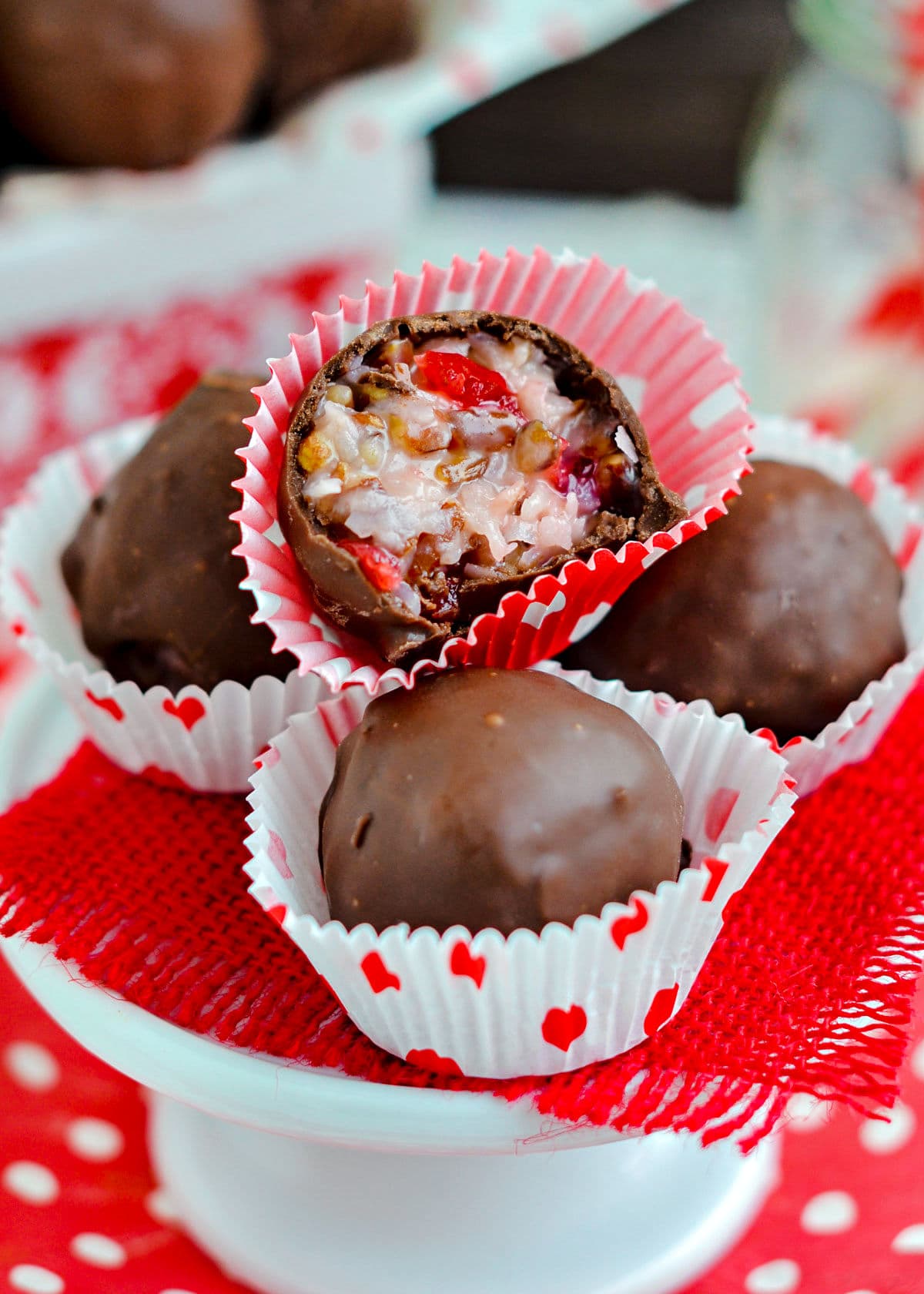 four candies sitting on small cupcake stand with red burlap beneath them. top candy is cut in half to show coconut and cherry intererior.