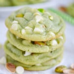 stack of four pistachio pudding cookies on white parchment studded with white chocolate chips and chopped pistachios.