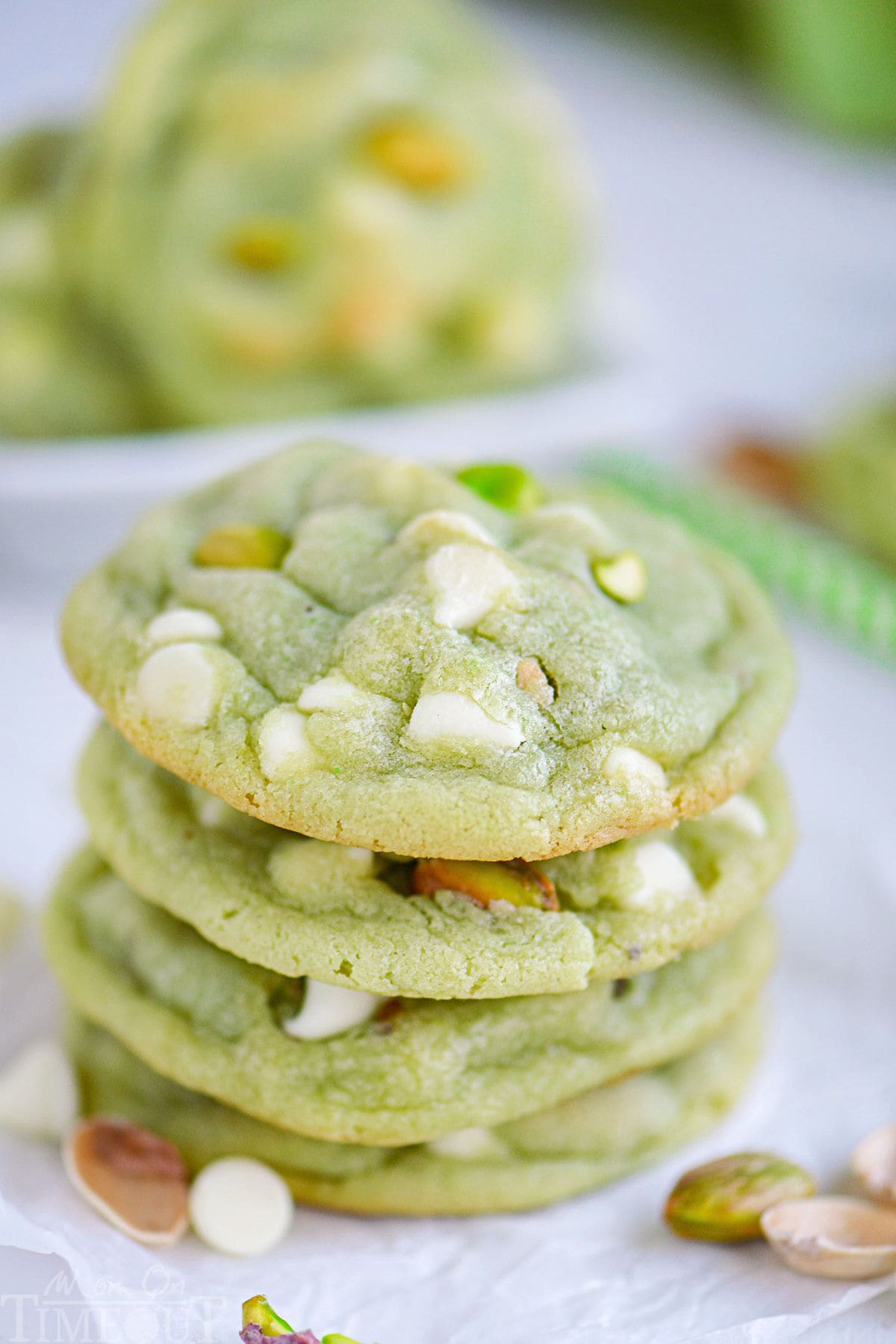 stack of four pistachio pudding cookies on white parchment studded with white chocolate chips and chopped pistachios.