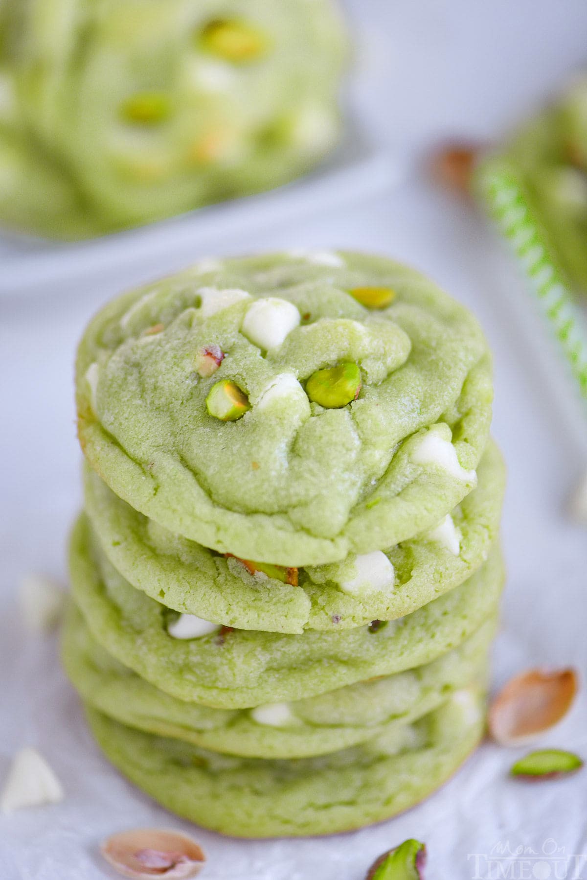 stack of five pistachio cookies on white parchment paper. the cookies have white chocolate chips and pistachios in them.