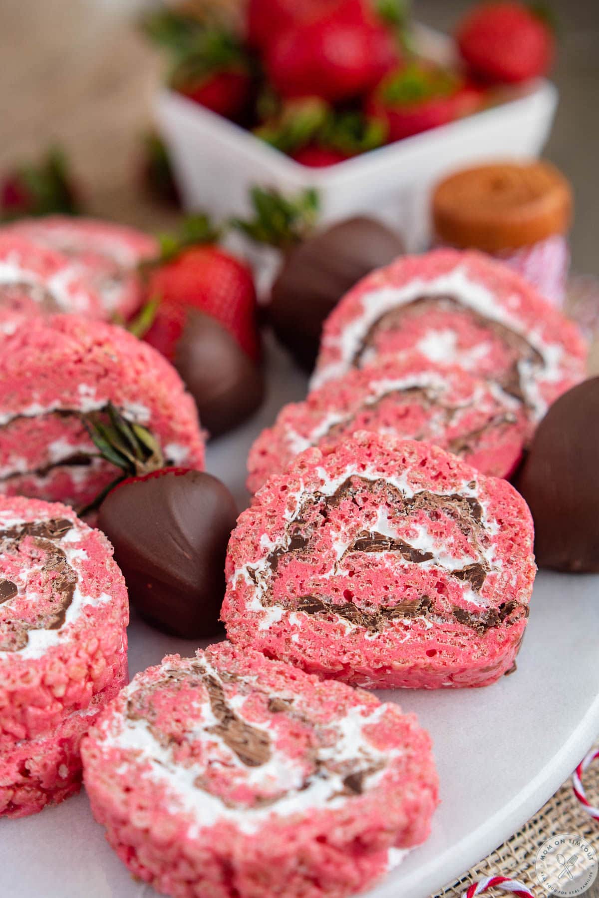 Strawberry chocolate rice krispie treat pinwheels on a white serving tray next to chocolate covered strawberries.