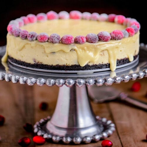 White chocolate cranberry cheesecake on silver cake stand. Decorated with sugared cranberries and topped with white chocolate ganache.