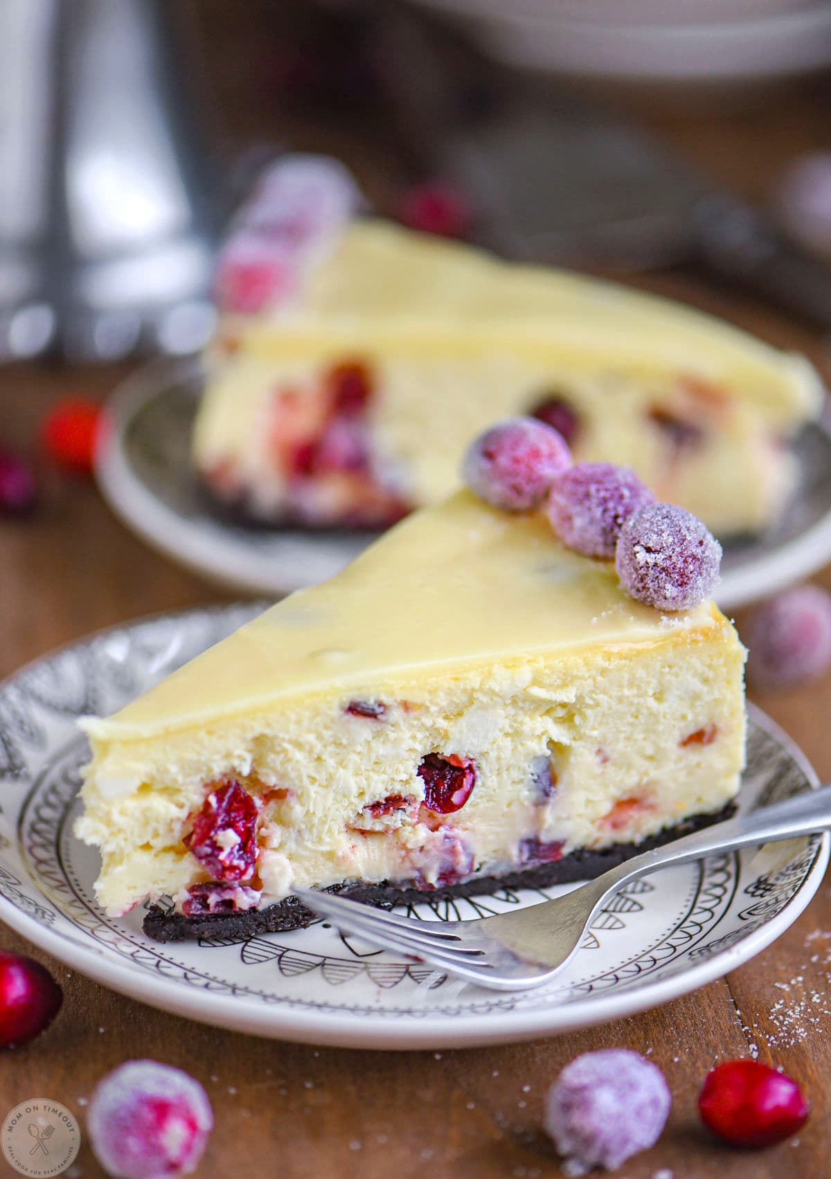 Slice of cranberry white chocolate cheesecake on round white plate. Sugared cranberries and white chocolate ganache on top of the cheesecake.