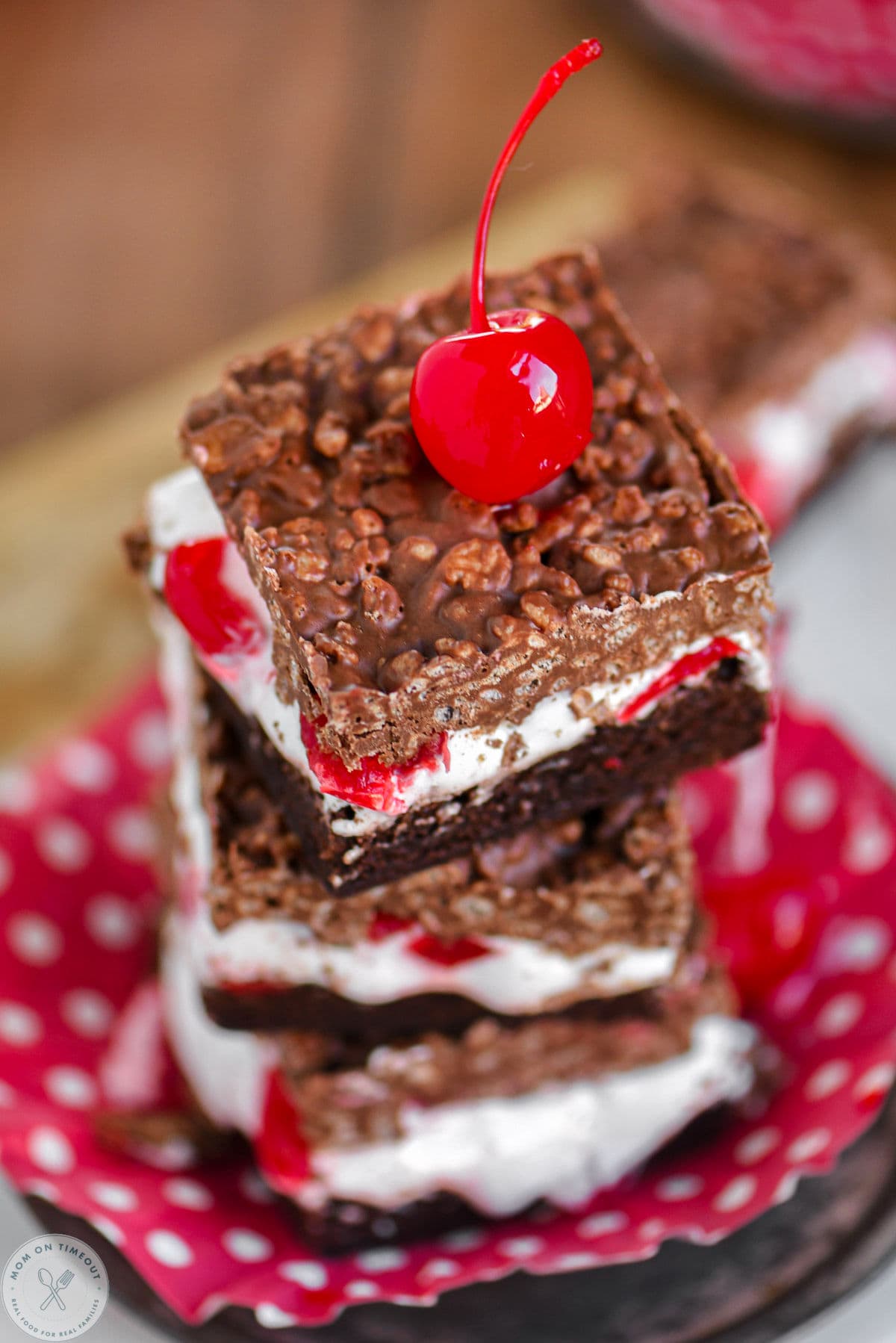 Angle overhead shot of cherry brownie marshmallow bars with crispy peanut butter chocolate topping. Bars are sitting on cupcake linerr that is red with white dots.