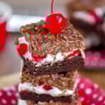 Stack of three cherry brownie dream bars on a metal plate with a maraschino cherry on top.