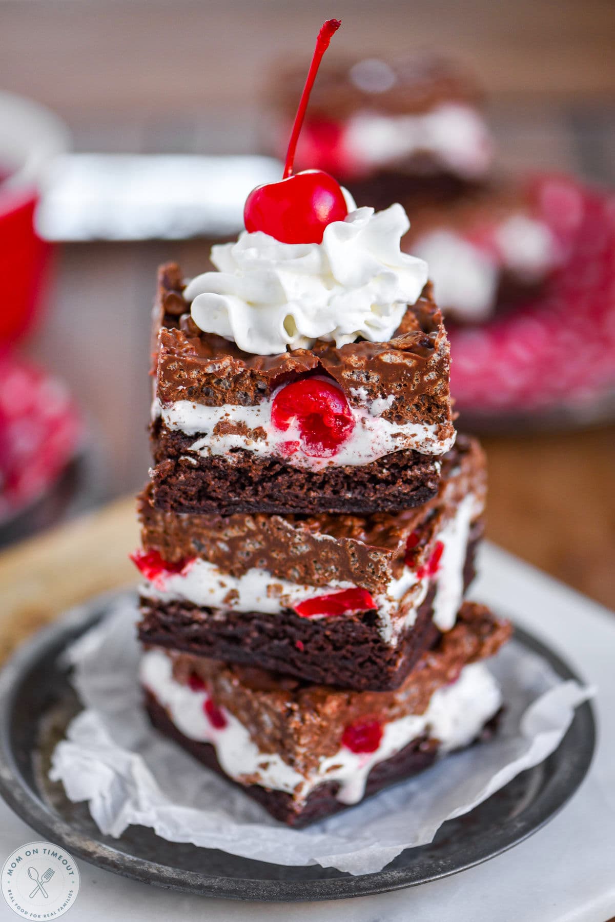 Three cherry dream bars stacked on parchment lined metal round plate. The brownies are topped with whipped cream and a maraschino cherry.