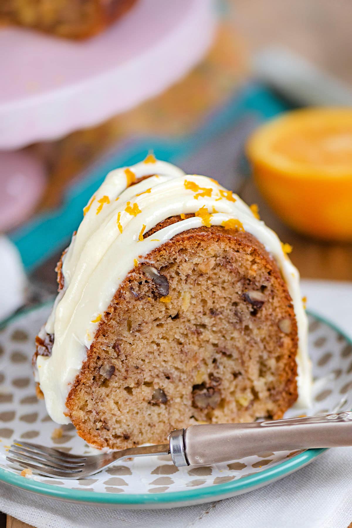 piece of hummingbird bundt cake on a small dessert plate sitting in front of a pink cake stand. the cake has been topped with a cream cheese frosting.