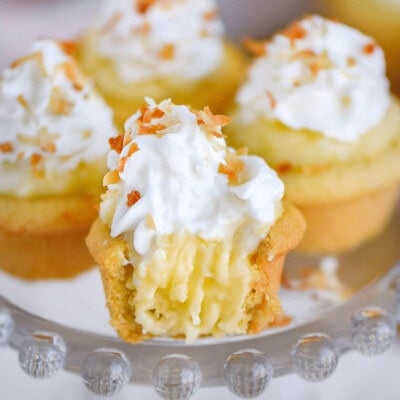 Four mini coconut cream pies on glass cake stand topped with whipped cream and toasted coconut. The front one has a bite taken out of it.