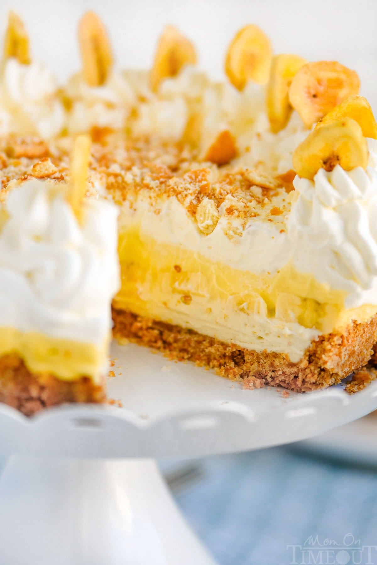 Shot of Banana Pudding Cheesecake Recipe on white metal scalloped cake stand with one piece cut and removed.