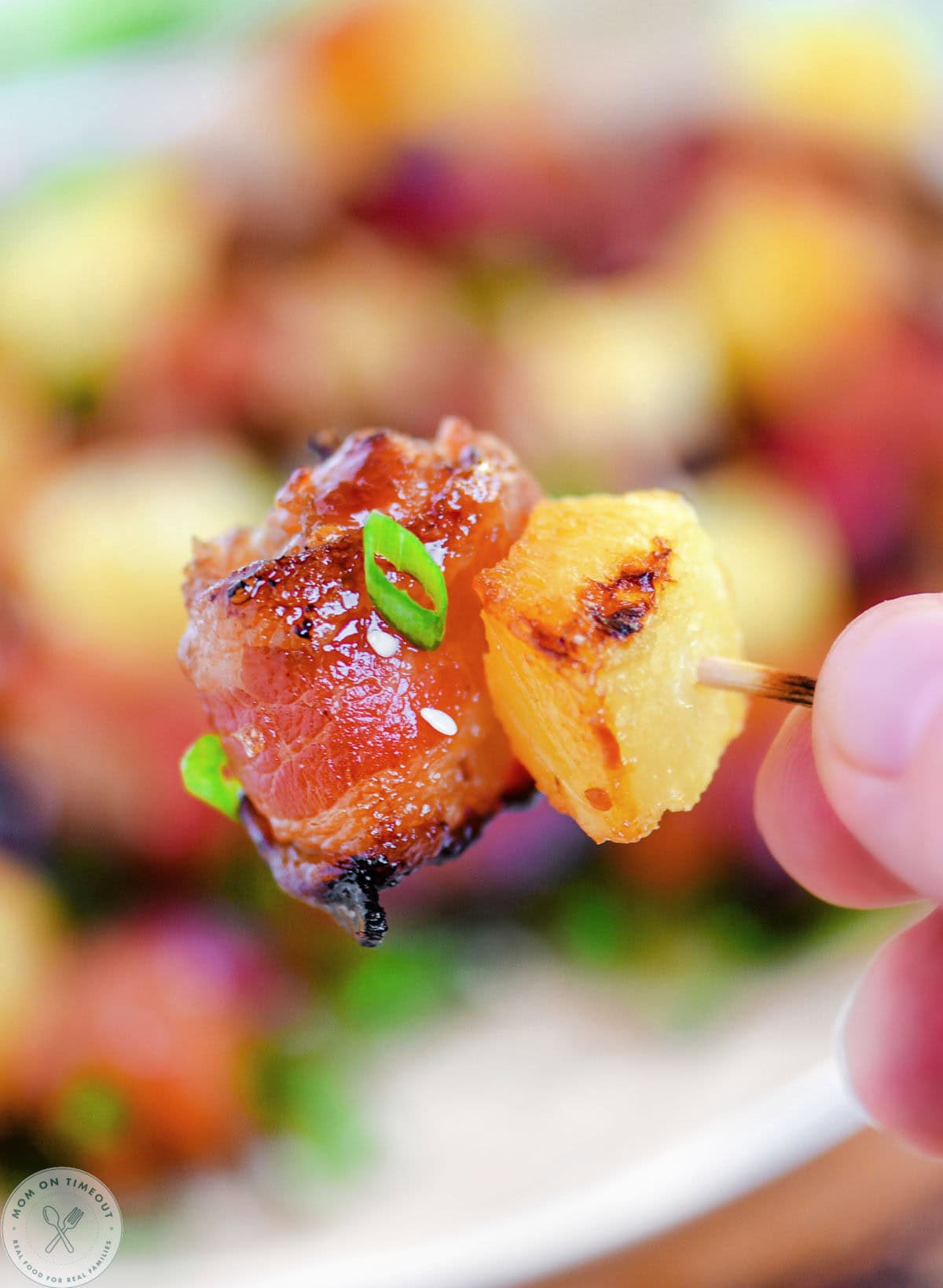 Bacon wrapped chicken teriyaki bite with pineapple on toothpick held up over platter full of the appetizer.