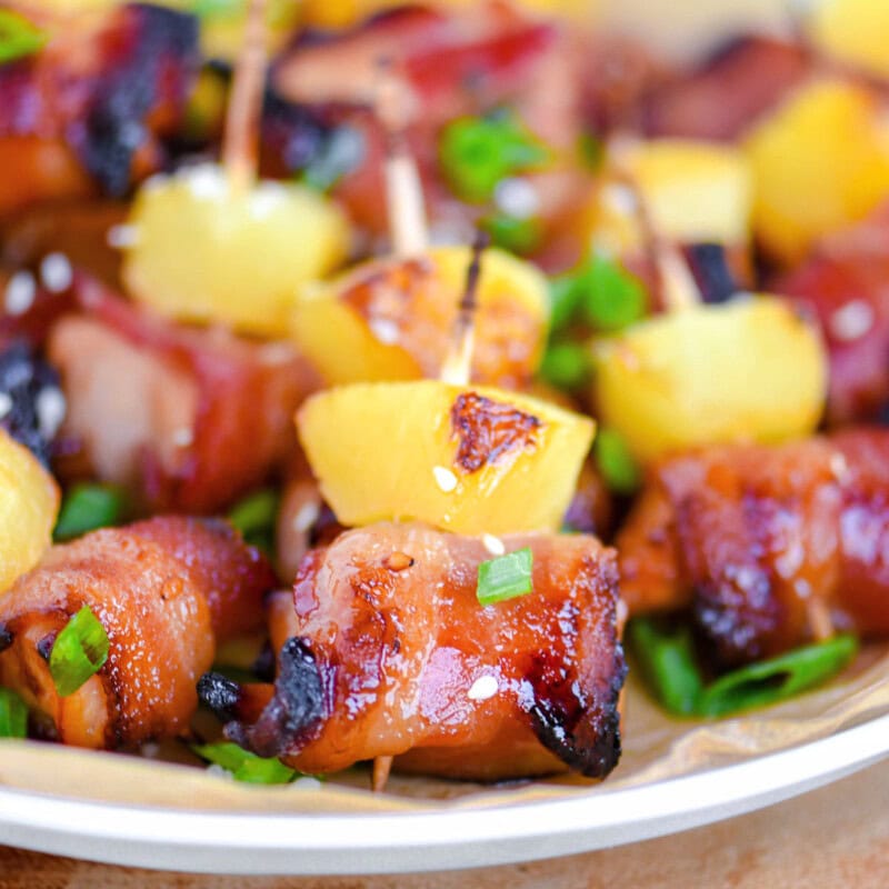 Plate of bacon wrapped chicken teriyaki bites with pineapple. Each bite is skewered with a toothpick and topped with green onions and sesame seeds.