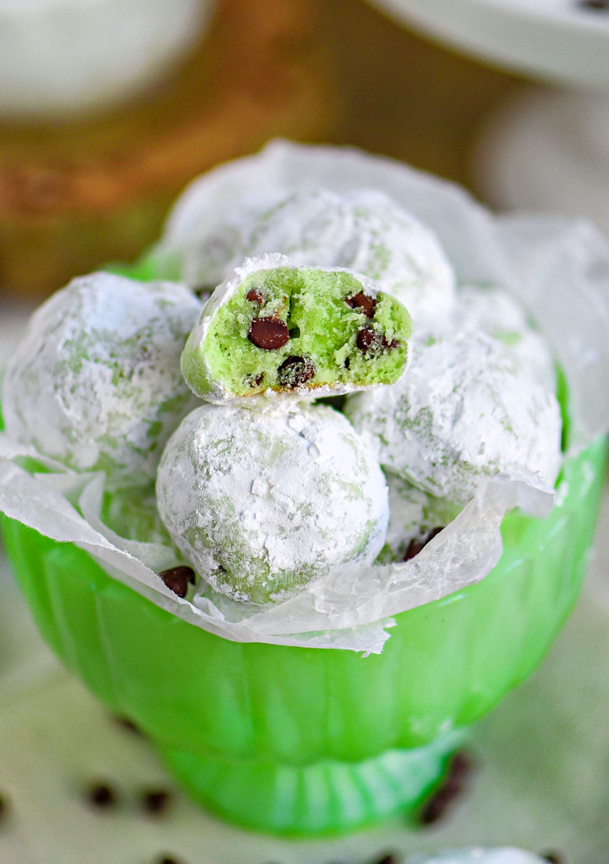 mint chocolate chip snowball cookies in a small green glass pedestal bowl lined with wax paper. One cookie is broken in half to show the green interior and mini chocolate chips.