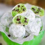 mint chocolate chip snowball cookies in a small green glass pedestal bowl lined with wax paper. A few cookies are broken in half to show the green interior and mini chocolate chips.