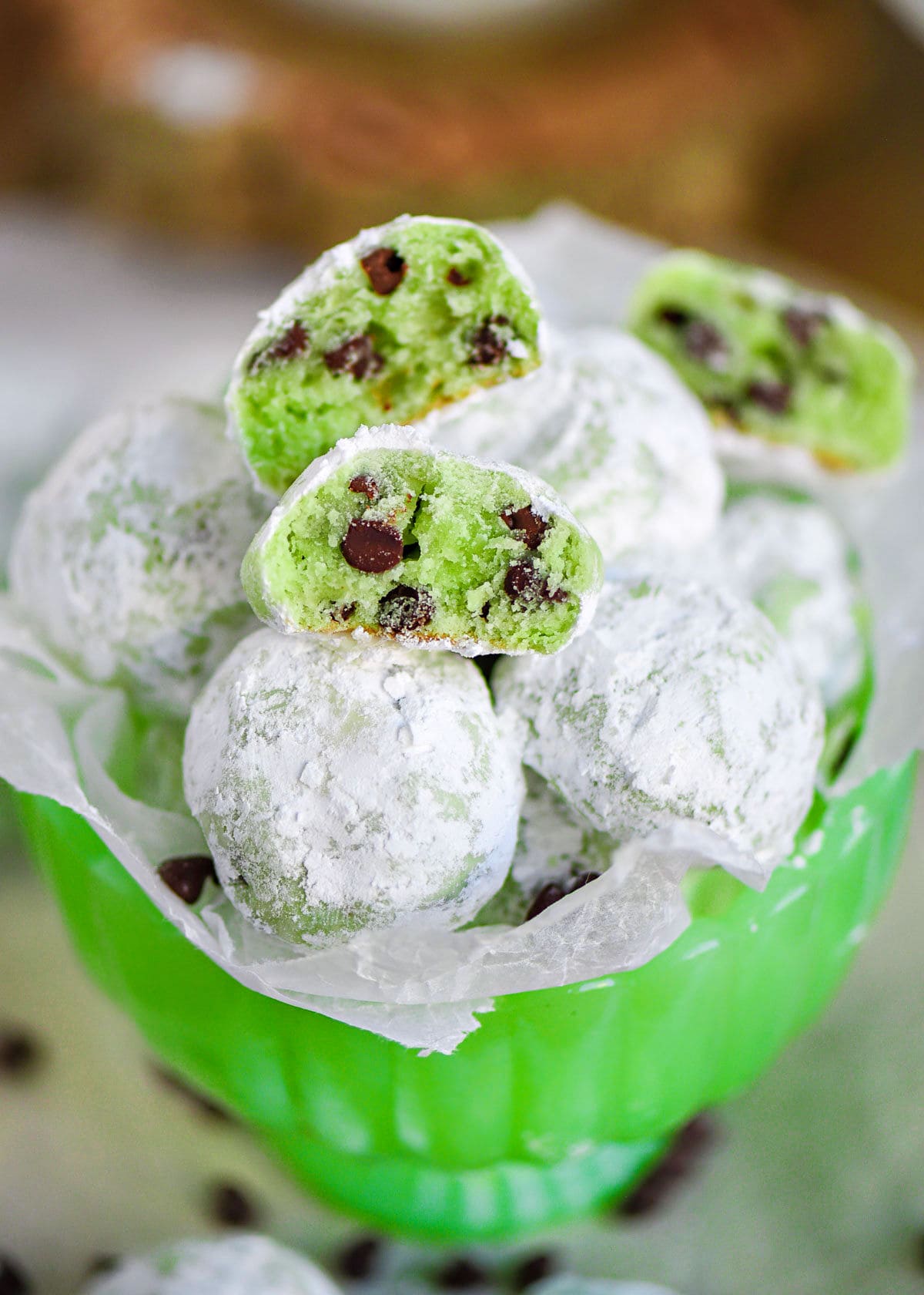 mint chocolate chip snowball cookies in a small green glass pedestal bowl lined with wax paper. A few cookies are broken in half to show the green interior and mini chocolate chips.