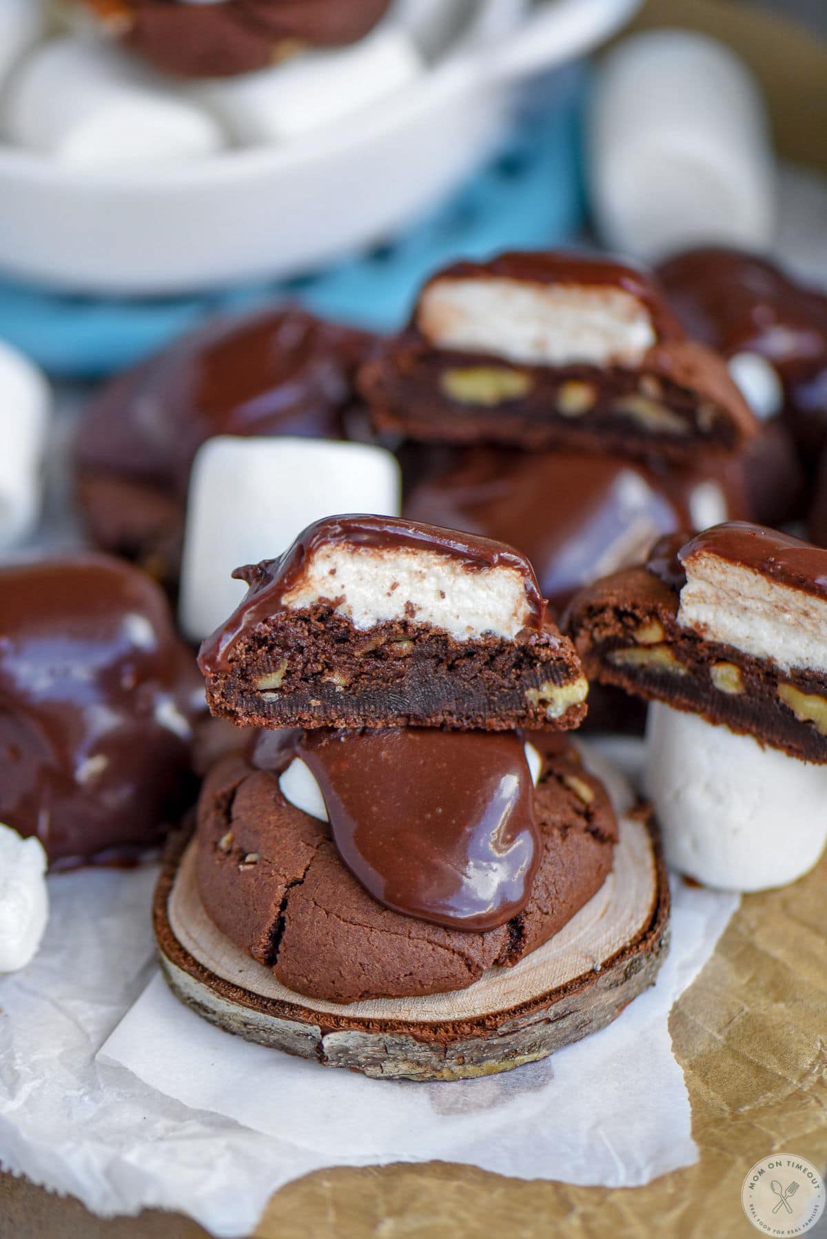 Chocolate marshmallow cookie recipe topped with fudge frosting sitting on wood coaster. More cookies cut in half can be seen in the background.