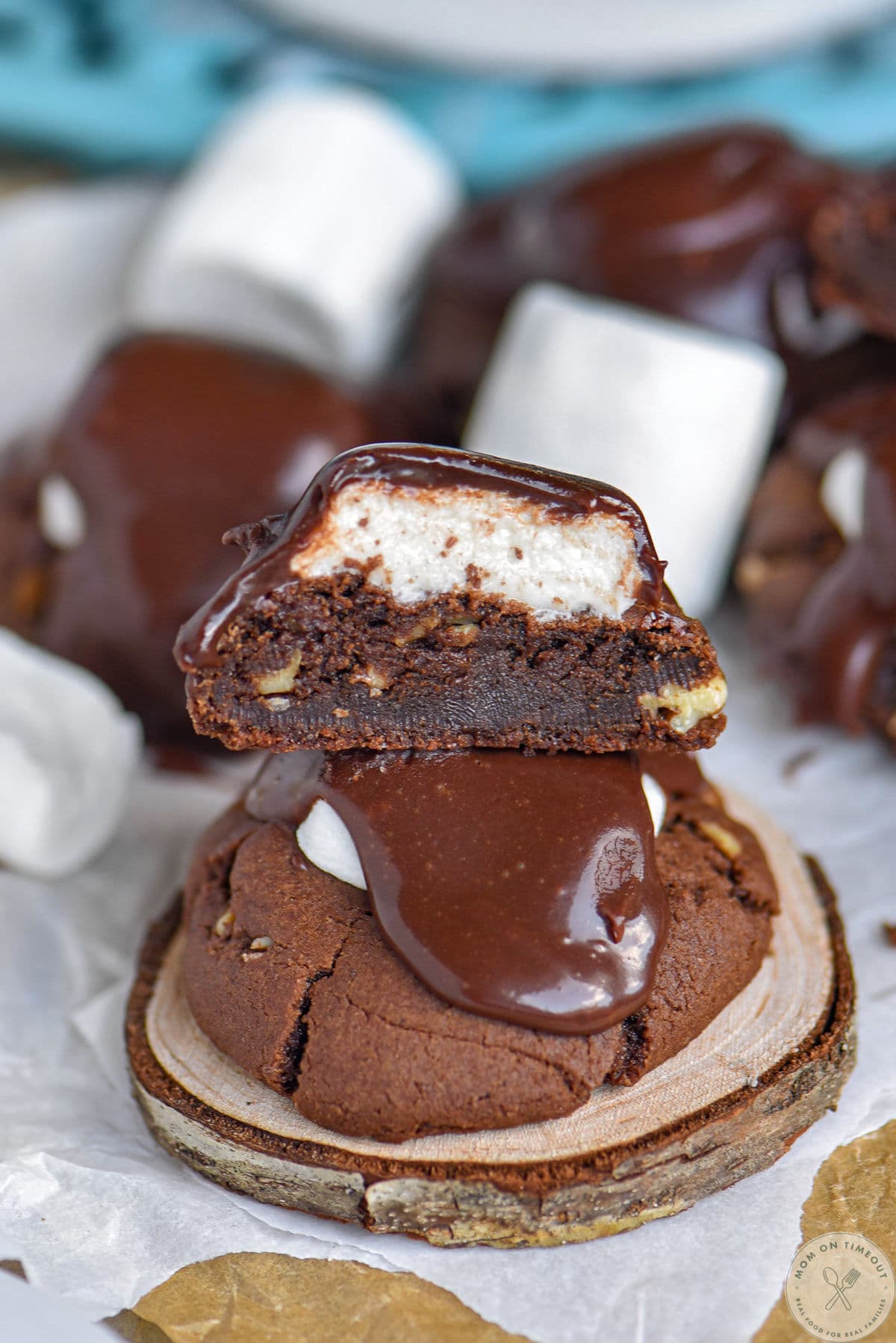Chocolate marshmallow cookies topped with fudge frosting sitting on wood coaster. More cookies cut in half can be seen in the background.